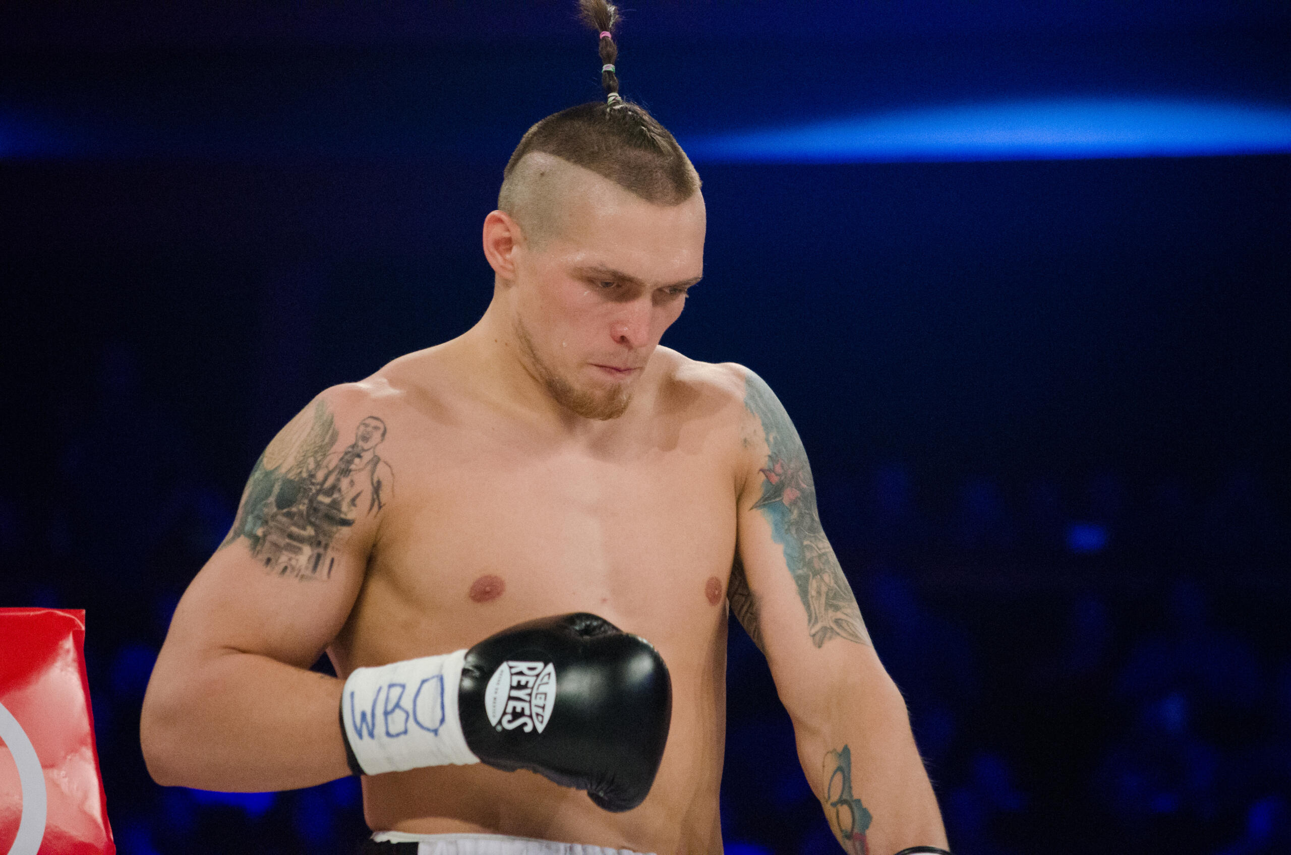 Un boxeador sin camiseta, con un característico peinado mohicano y tatuajes en los brazos, que lleva guantes de boxeo negros de la marca Cleto Reyes con las siglas «WBO» escritas en ellos.